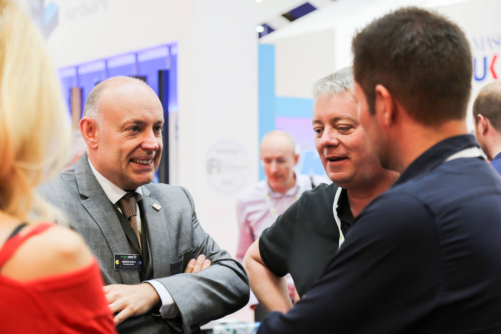 Andrew Scott, wearing a grey suit, interacts with colleagues during a business event, exemplifying his role as a leader and entrepreneur.