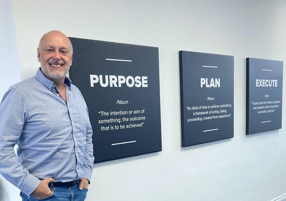 Andrew Scott, smiling in a casual shirt, stands next to Purplex Marketing's "Purpose, Plan, Execute" wall, reflecting the company's core strategic values.