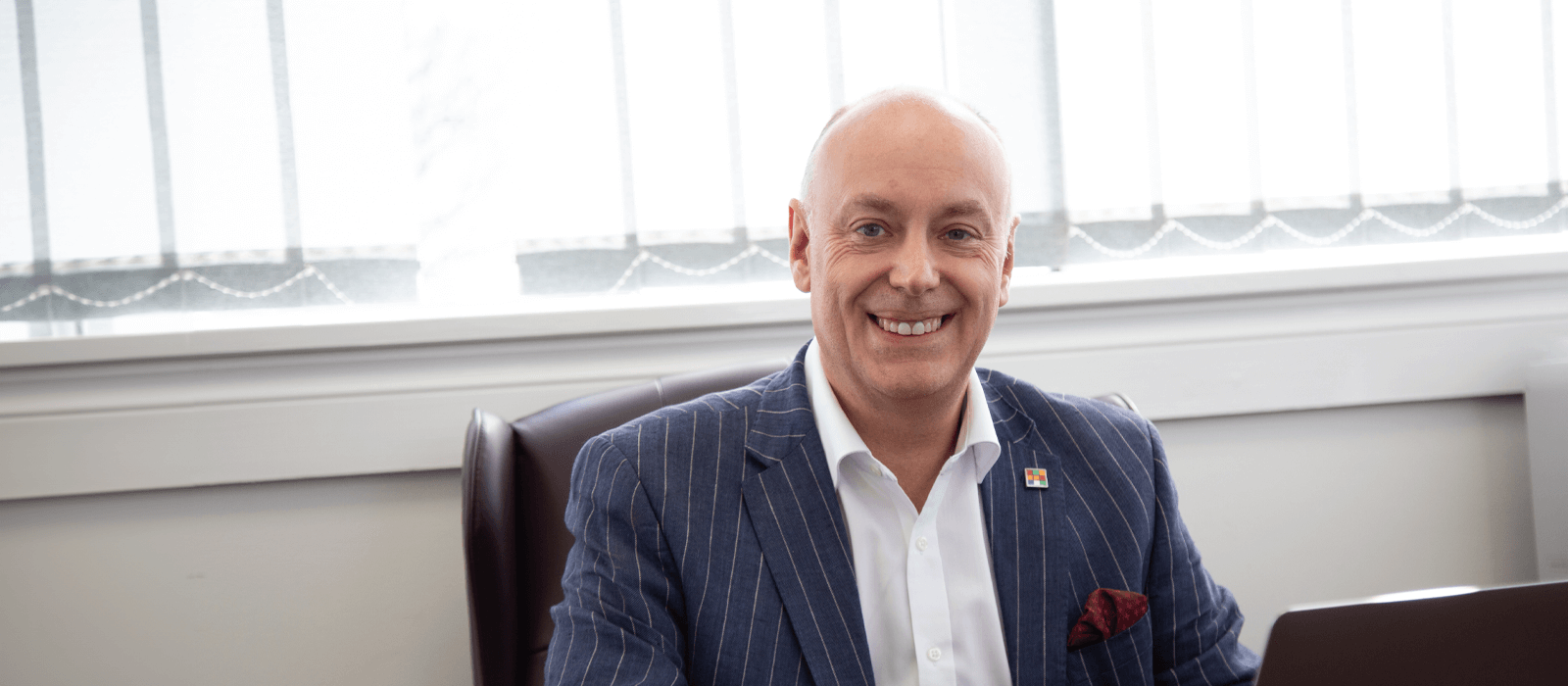 Entrepreneur Andrew Scott, dressed in a pinstripe blazer, smiles at his desk at the Ascot Group, after being appointed to the Forbes Business Council.