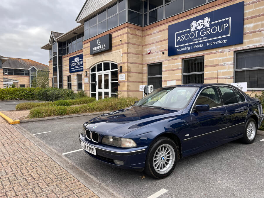 A blue BMW is parked outside the Ascot Group headquarters, with the building’s signage visible. The image connects the car’s symbolic role in Andrew Scott’s journey to the success of the company he built.